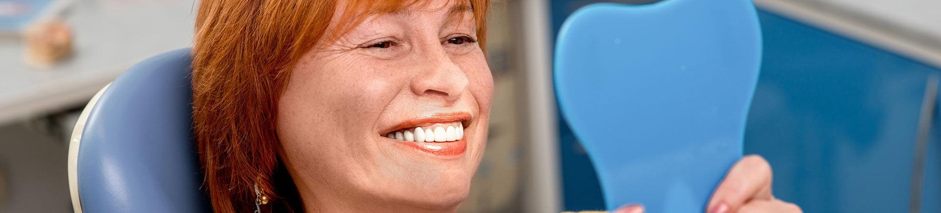 Happy mature woman in a dental chair looking at her perfect teeth in a mirror.