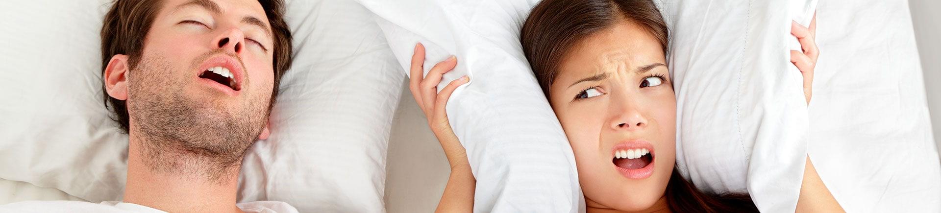 A snoring man sleeping next to a frustrated woman covering her ears with a pillow.