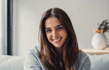 A young woman with long brown hair sits indoors, smiling at the camera. A white vase with dried flowers is visible in the background.