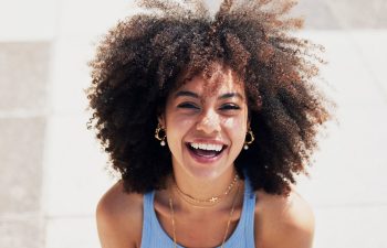 A person with curly hair smiles broadly while looking up at the camera, wearing a light blue top and gold jewelry, outdoors in bright natural light.