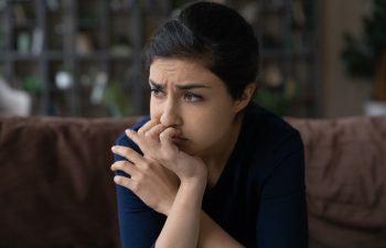A woman with dark hair sits on a couch, resting her chin on her hand and looking to the side with a concerned expression.