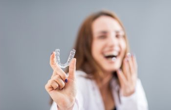A person holds up a clear dental aligner with one hand while smiling, with their face out of focus in the background.